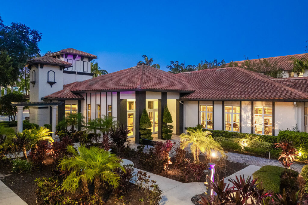 A modern Mediterranean-style building with red-tiled roof, large windows, and lush landscaping, illuminated by outdoor lights at dusk. Palm trees and tropical plants line the walkway in the foreground.